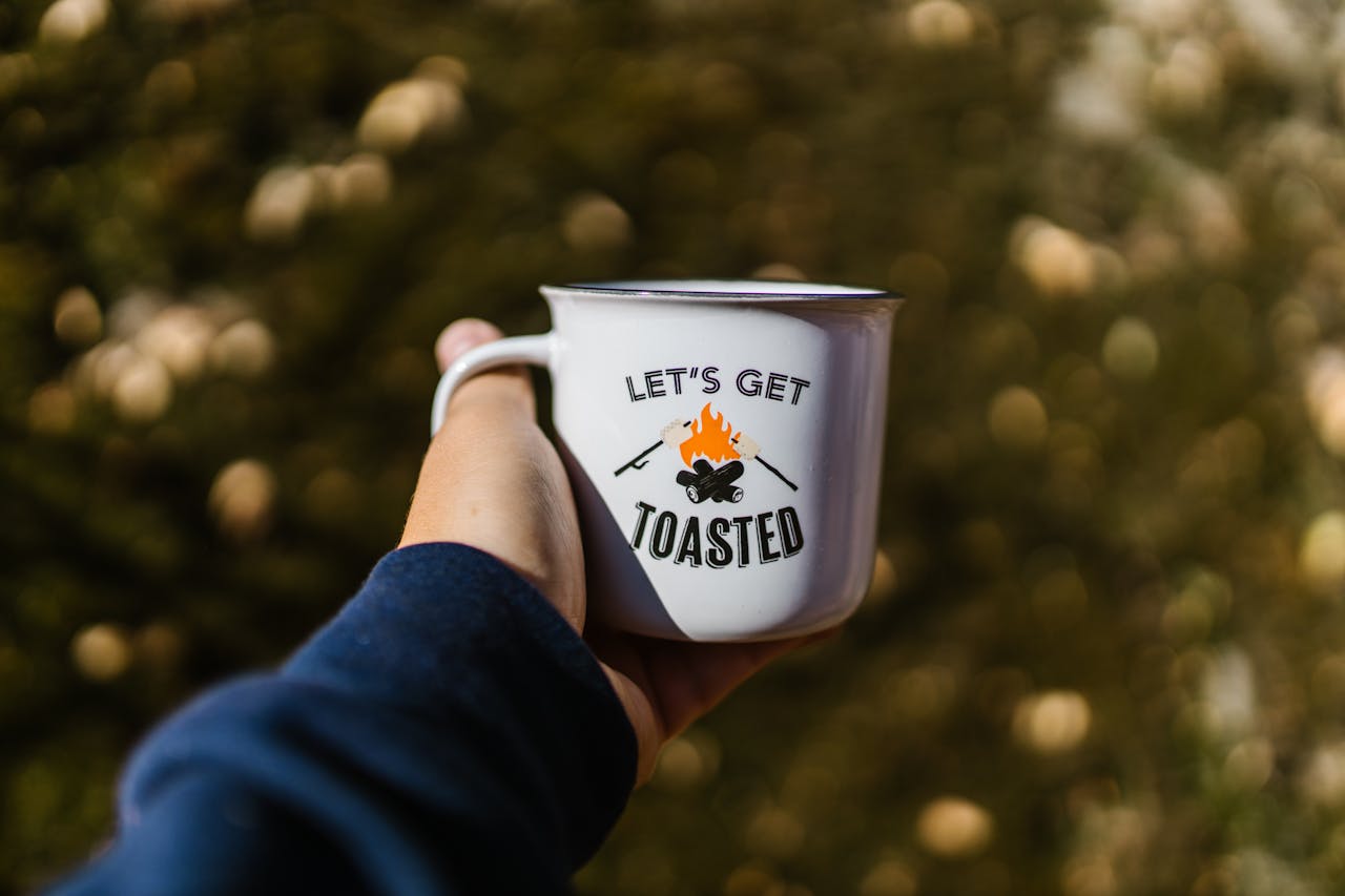 Person holding enamel mug with 'Let's Get Toasted' design, ideal for camping vibes.